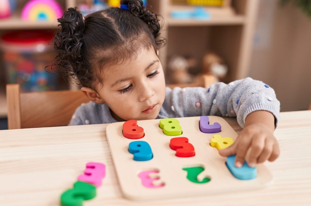 Adorable hispanic girl playing with maths puzzle game sitting on table at kindergarten